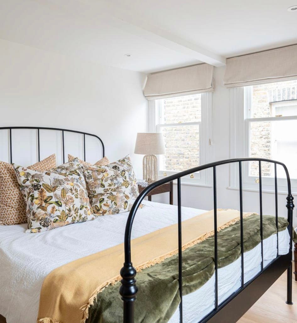 Guest bedroom with a black iron bedstead, linen roman blinds and botanical-print cushions